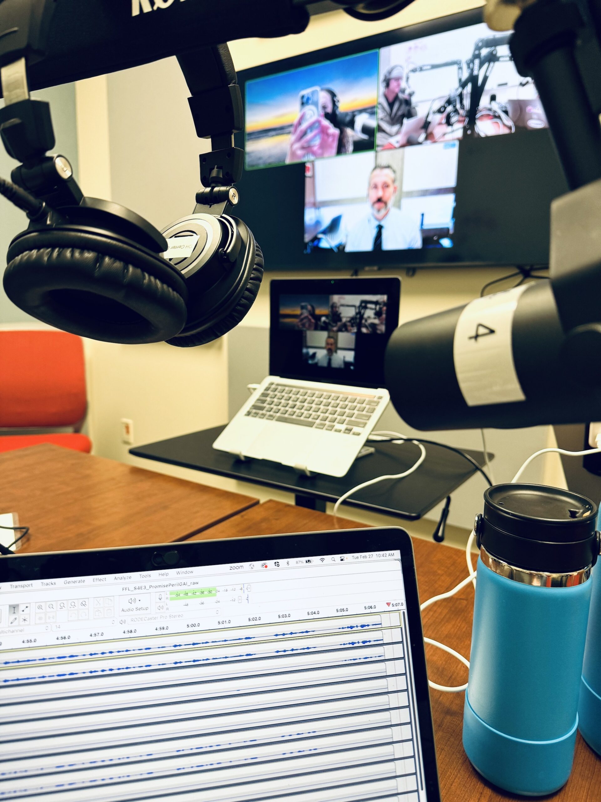  podcast gear in the foreground with guests appearing via Zoom on a TV screen in the background